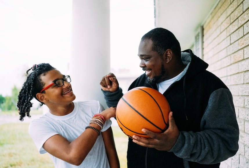 A smiling adult holding a basketball playfully interacts with a child wearing glasses and a white t-shirt. The two are standing outside near a building, enjoying a lighthearted moment. The child is laughing while looking up at the adult, who seems to be giving some basketball tips.